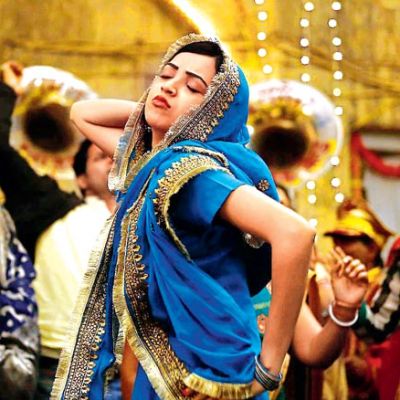 Still from a Hindi film 'Lipstick under my Burkha'. A young woman wearing blue-coloured lehnga is dancing merrily in an Indian wedding. She has one hand on her hip, another behind her head, and is tilting upwards with her eyes closed, and lips pout.