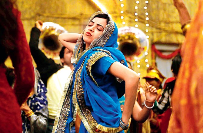 Still from a Hindi film 'Lipstick under my Burkha'. A young woman wearing blue-coloured lehnga is dancing merrily in an Indian wedding. She has one hand on her hip, another behind her head, and is tilting upwards with her eyes closed, and lips pout.