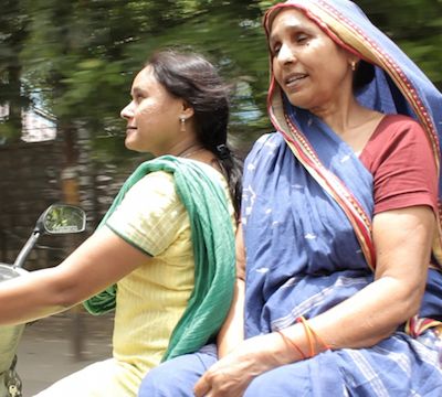 Two women riding a scooter. A younger Indian woman wearing a low pony-tail and a suit is on the wheel, with an older woman who is wearing a saree with which she covers her head, is sitting at the back facing sideways.