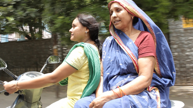 Two women riding a scooter. A younger Indian woman wearing a low pony-tail and a suit is on the wheel, with an older woman who is wearing a saree with which she covers her head, is sitting at the back facing sideways.