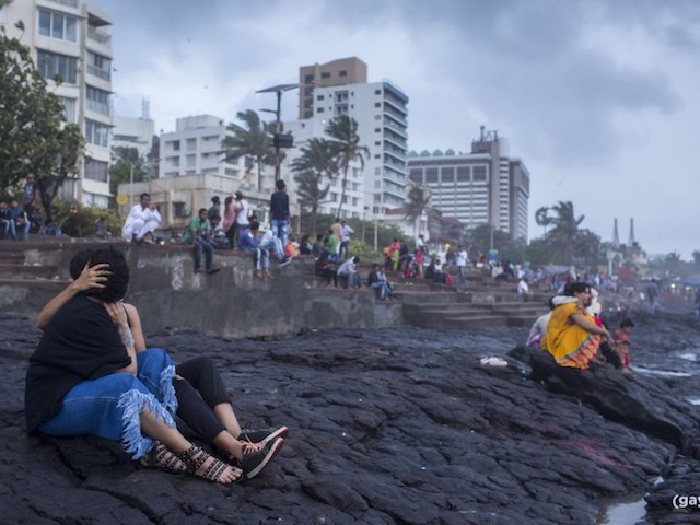 Couples sitting and kissing by a beach-side of an industrailised smoggy area, with palm trees and its' leaves waving in a strong wind.