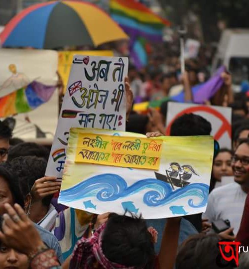 Photo of a vibrant queer pride march. There is a huge crowd, lots of rainbow-coloured umbrellas, flags, drawings, and writings on placards. One placard reads "ab toh angrej bhi maan gaye" in Hindi.