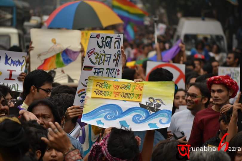 Photo of a vibrant queer pride march. There is a huge crowd, lots of rainbow-coloured umbrellas, flags, drawings, and writings on placards. One placard reads "ab toh angrej bhi maan gaye" in Hindi.