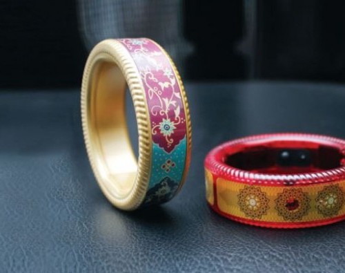 Two thick bangles are placed on a table. One is balanced in a vertical position as if to roll, the other is placed horizontally. One has red boundary, with thick yellow strip of colour in the middle, and a circular design in golden colour. The other one has golden boundary, and has traditional Indian design in blue and red in the middle.