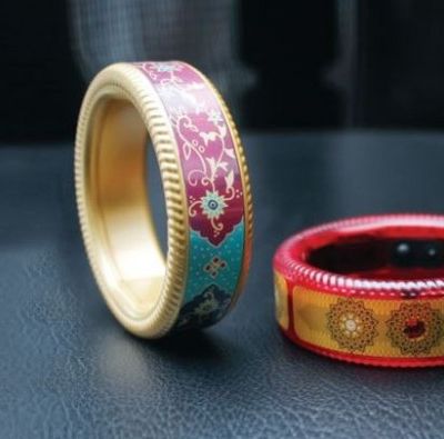 Two thick bangles are placed on a table. One is balanced in a vertical position as if to roll, the other is placed horizontally. One has red boundary, with thick yellow strip of colour in the middle, and a circular design in golden colour. The other one has golden boundary, and has traditional Indian design in blue and red in the middle.