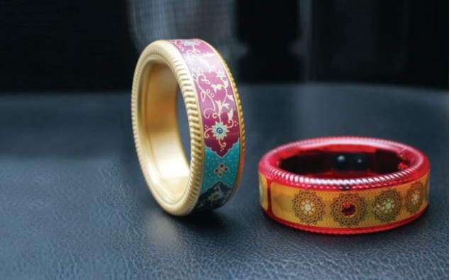 Two thick bangles are placed on a table. One is balanced in a vertical position as if to roll, the other is placed horizontally. One has red boundary, with thick yellow strip of colour in the middle, and a circular design in golden colour. The other one has golden boundary, and has traditional Indian design in blue and red in the middle.