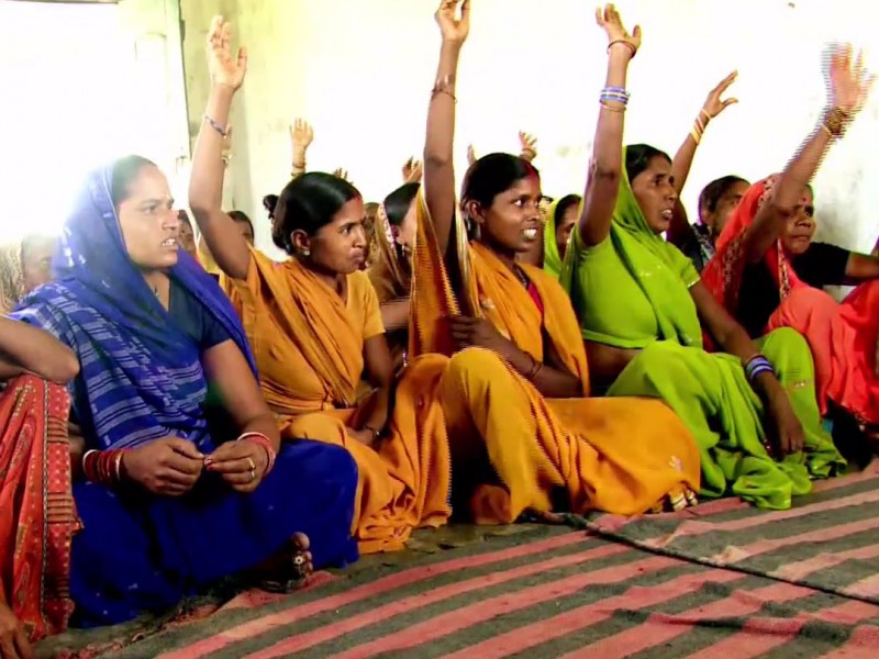 Several young Indian rural women sitting in a row on a carpet. Some have raised their hands, as if in answer to a question. They are wearing bright-coloured sarees, and bangles. Some are wearing bindis too.