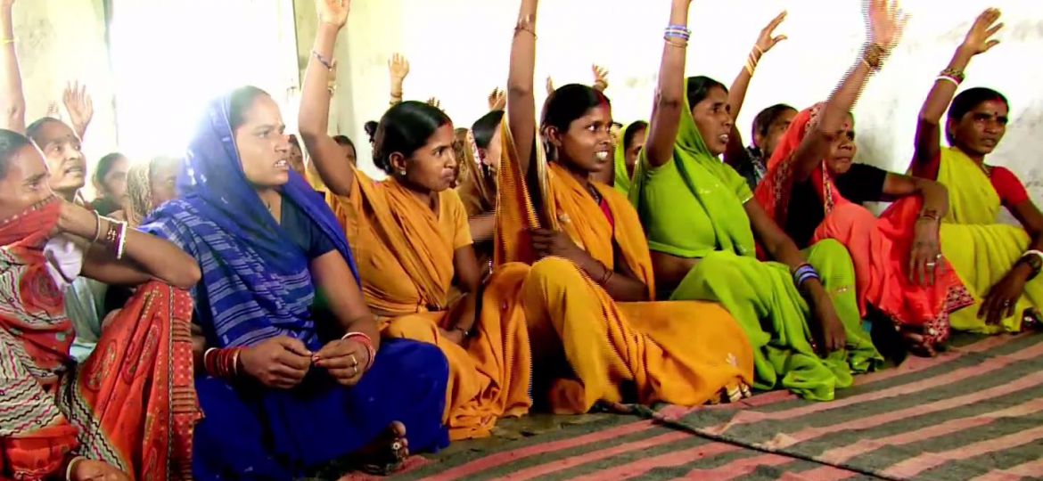 Several young Indian rural women sitting in a row on a carpet. Some have raised their hands, as if in answer to a question. They are wearing bright-coloured sarees, and bangles. Some are wearing bindis too.