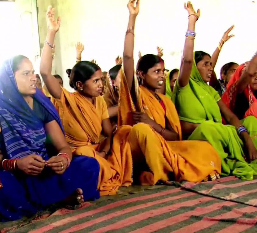 Several young Indian rural women sitting in a row on a carpet. Some have raised their hands, as if in answer to a question. They are wearing bright-coloured sarees, and bangles. Some are wearing bindis too.