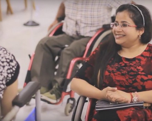 A woman in a wheelchair, smiling, conversing with another woman who is also on a wheelchair. She has long hair pulled to one side, is wearing frameless specks, and is wearing a red and black top.