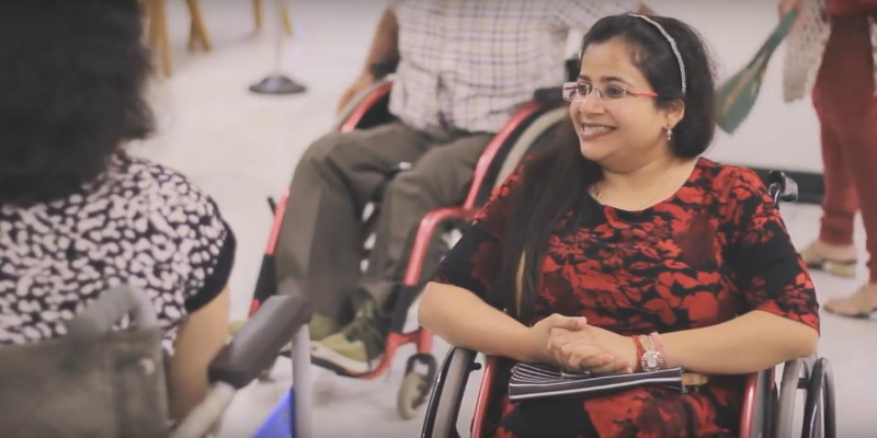 A woman in a wheelchair, smiling, conversing with another woman who is also on a wheelchair. She has long hair pulled to one side, is wearing frameless specks, and is wearing a red and black top.