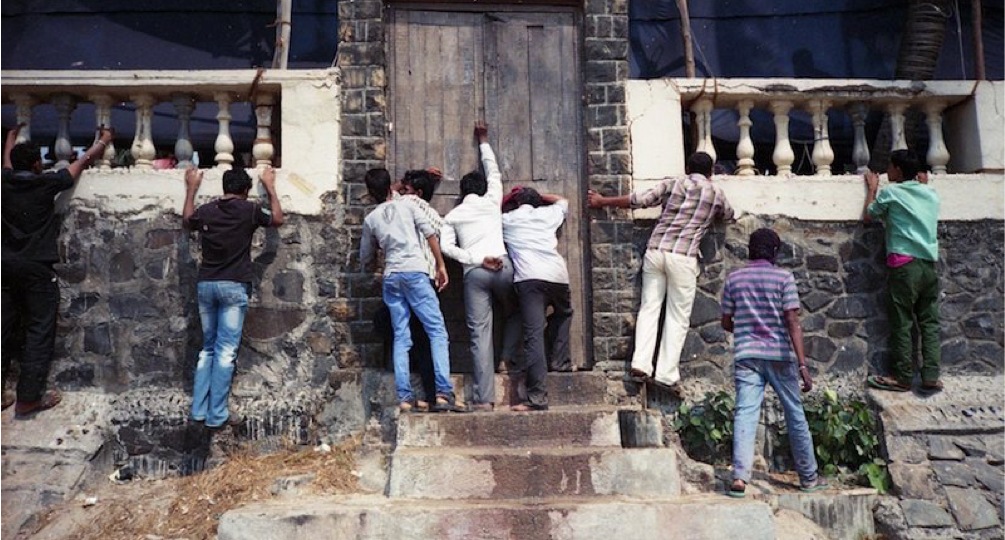 A few men knocking the door of an old Indian house. A few other men are trying to peek into the house through a very low railing.