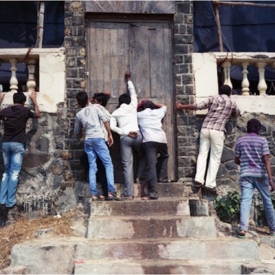 A few men knocking the door of an old Indian house. A few other men are trying to peek into the house through a very low railing.