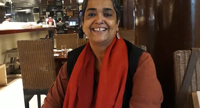 Picture of feminist activist Pramada Menon, she ihas short hair and is wearing a maroon kurta, black waistcoat and a red dupatta