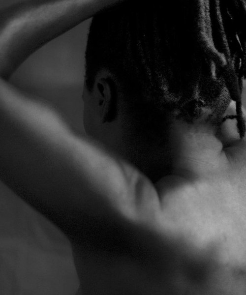 A black and white photo of a naked woman with her back turned to us. Her hands go up her head, forming a kind of band for her Senegalese twists-styled hair that fall in a kind of pony tail till the top of her neck.