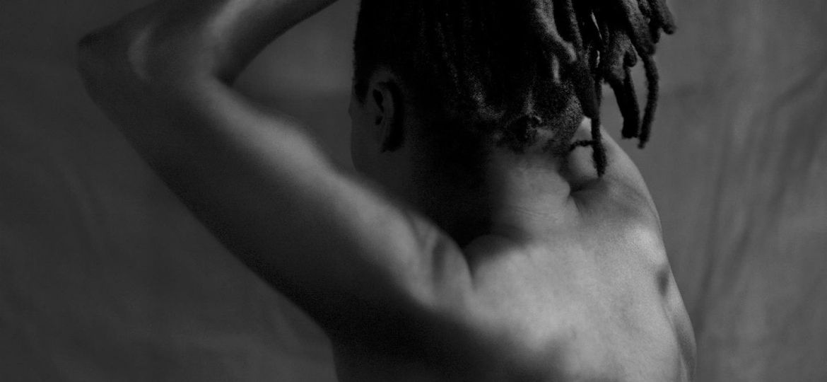 A black and white photo of a naked woman with her back turned to us. Her hands go up her head, forming a kind of band for her Senegalese twists-styled hair that fall in a kind of pony tail till the top of her neck.