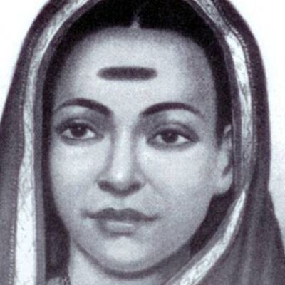 A black-and-white headshot of Savitribai Phule. She has covered her head with her saree, which has a white border. She is looking straight, expression-less. She has a big horizontal mark right at the centre of her forehead.