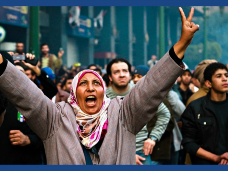 On a foggy winter morning, a woman protestor waves a victory sign, looks upwards, and shouts what could be possibly be a slogan. She wears a pink and white coloured headscarf and a grey coat. There are hoards of other protestors in the background too.