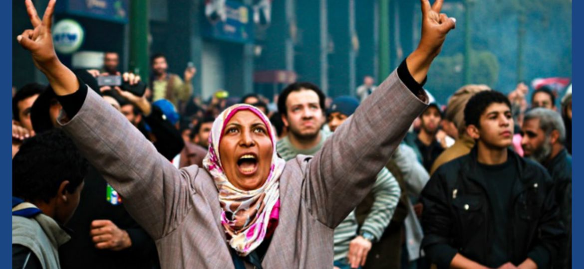 On a foggy winter morning, a woman protestor waves a victory sign, looks upwards, and shouts what could be possibly be a slogan. She wears a pink and white coloured headscarf and a grey coat. There are hoards of other protestors in the background too.