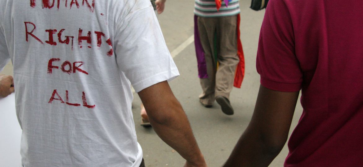 Photo from a queer pride march. We can see the back of two men holding hands. On the back of one's tee shirt is witten "Human rights for all" in caps by a red marker. The photo is cropped such that we can see only their torso and hands. In the background we can see more people walking in the march.