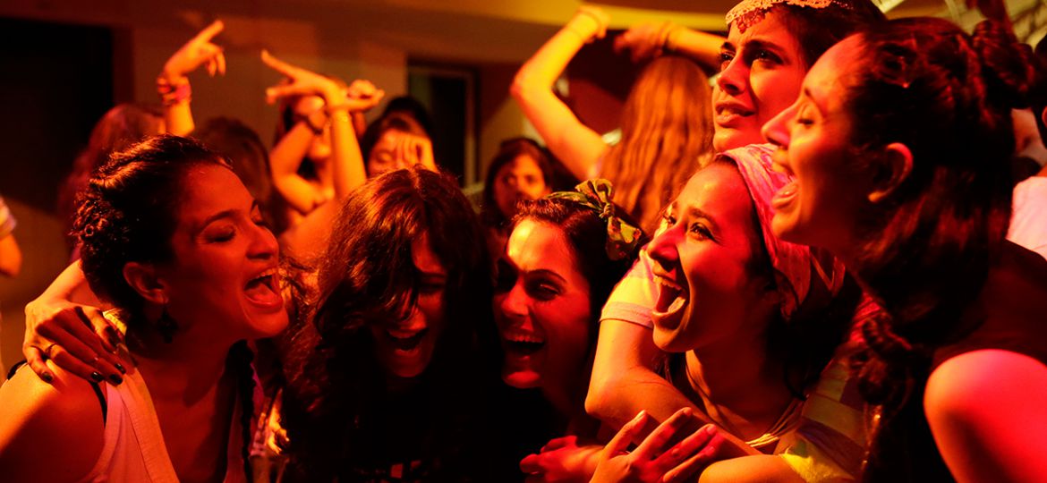 Scene from a Hindi movie 'Angry Indian Goddesses'. The six protagonists are standing together laughing, in a disco with bright lighting.
