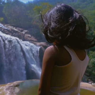 एक बच्ची (जिसकी पीठ और बाल दिख रखे है) पानी के झरने की और देख रही है, झरने क आस-पास हरियाली और पहाड़ है। बच्ची के बाल कंधे तक है और उसने हलके भूरे रंग की कमीज़ पहनी है