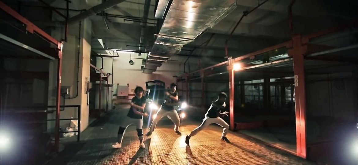 A still from a music video, showing a bunch of men dancing on the street.