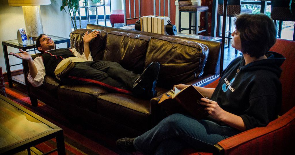 a man dressed in black trousers, white shirt, and black waistcoat, lies on a couch. his hands seem to be moving like he's making conversation. a woman sits on a couch beside him, she has a notebook in her hand as if she's listening to what he's saying and making notes.