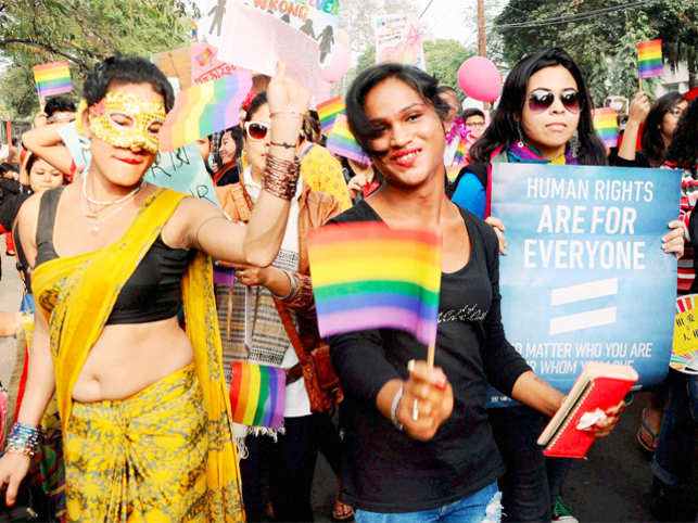 A picture from a pride parade; in the center, a woman in a black shirt holds up a pride flag, to her right, a trans woman in a bright orange saree wears a golden mask, and to her left, a woman in a green top holds up a placard with a slogan