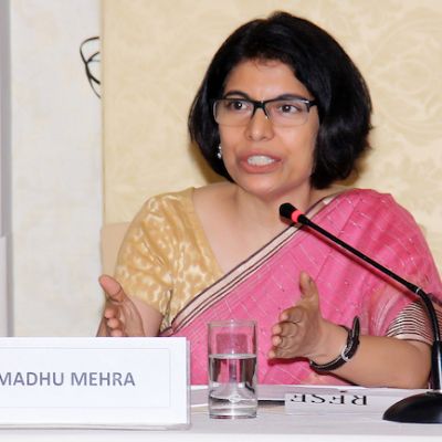 picture of lawyer and activist madhu mehra. she wears a pink saree with a beige blouse and sits at a desk before a microphone
