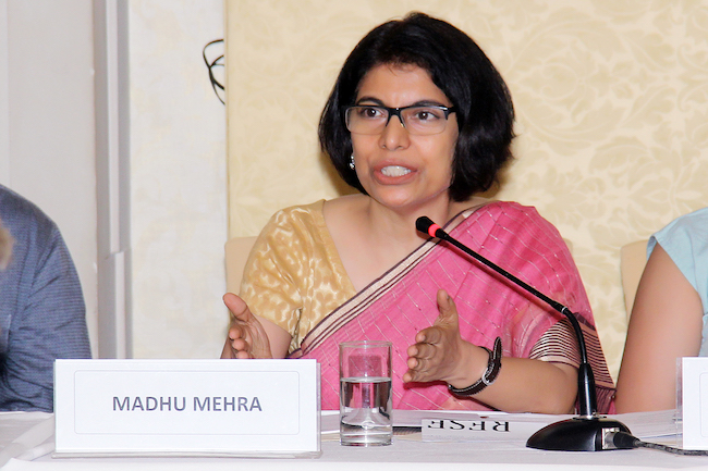 picture of lawyer and activist madhu mehra. she wears a pink saree with a beige blouse and sits at a desk before a microphone