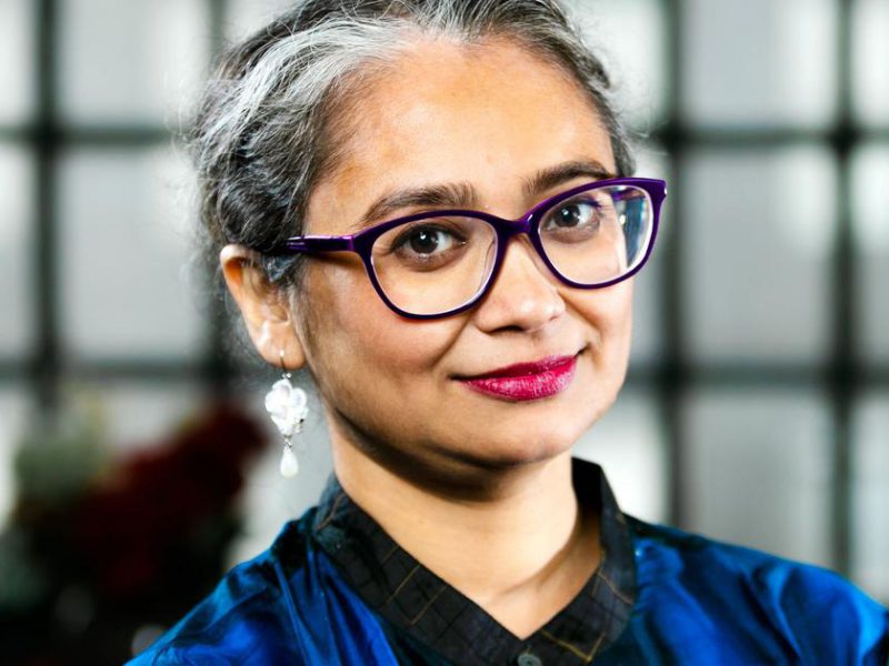 Picture of author Manjima Bhattacharya. She has short white-and-grey hair and is wearing dark thick-rimmed glasses