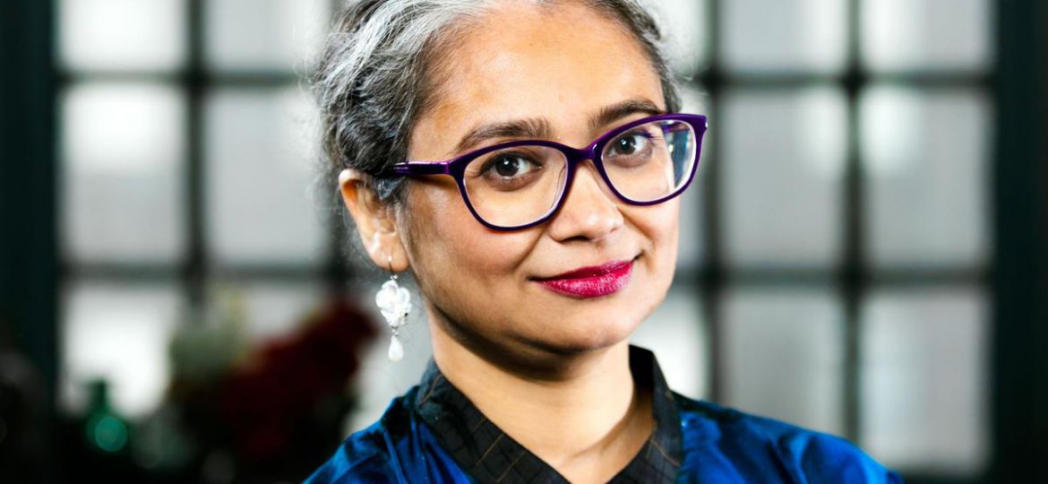 Picture of author Manjima Bhattacharya. She has short white-and-grey hair and is wearing dark thick-rimmed glasses