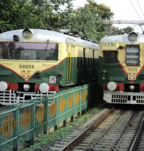 Picture of two trains, one already present on the platform, and another approaching the platform. They are both green in colour with off-white stripes on them