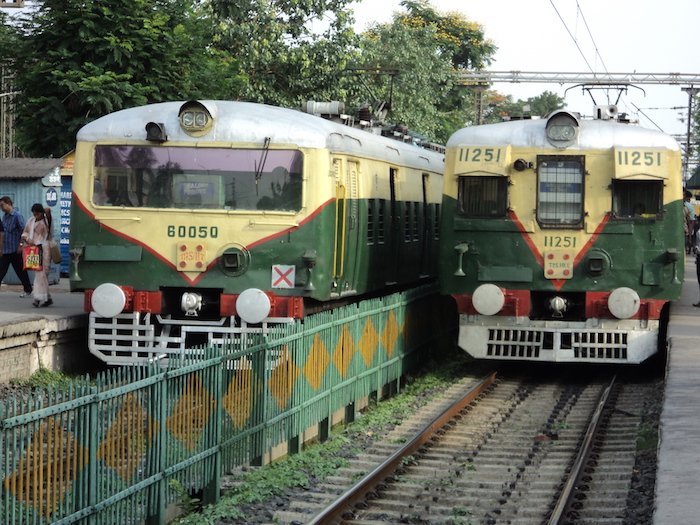 Picture of two trains, one already present on the platform, and another approaching the platform. They are both green in colour with off-white stripes on them