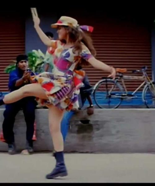 A still from the film Rangeela a girl in a white dress with floral patterns and a hat dances with one leg up