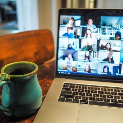 An image of a laptop and a cup held up close to it. A video conferencing platform is open on the laptop’s screen, divided into squares displaying the participants’ faces.