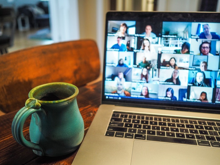 An image of a laptop and a cup held up close to it. A video conferencing platform is open on the laptop’s screen, divided into squares displaying the participants’ faces.