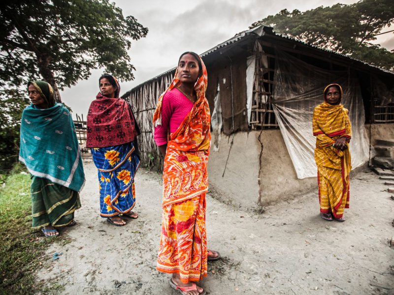 A photograph of four women, wearing sarees, standing in front of a hut. They are looking directly at the camera.
