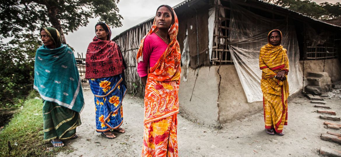 A photograph of four women, wearing sarees, standing in front of a hut. They are looking directly at the camera.