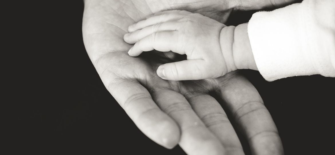 A black and white photograph of an infant’s hand resting on an older individual’s outstretched palm.