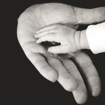 A black and white photograph of an infant’s hand resting on an older individual’s outstretched palm.