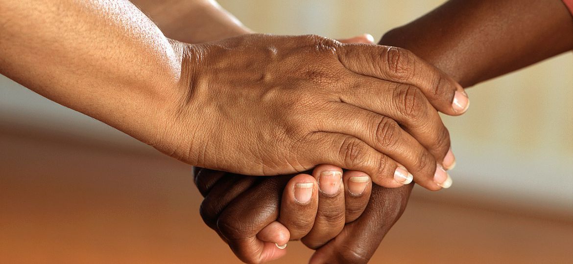 A photograph of two individual’s hands, from their forearms. One of the individuals is clasping the others’ hand in theirs.