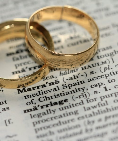 Two golden-coloured band rings atop a dictionary page zoomed in on the word ‘marriage’. Above that, the definition of the word ‘marrano’.