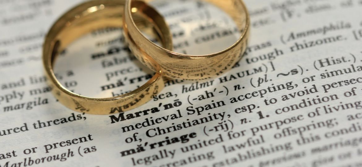 Two golden-coloured band rings atop a dictionary page zoomed in on the word ‘marriage’. Above that, the definition of the word ‘marrano’.