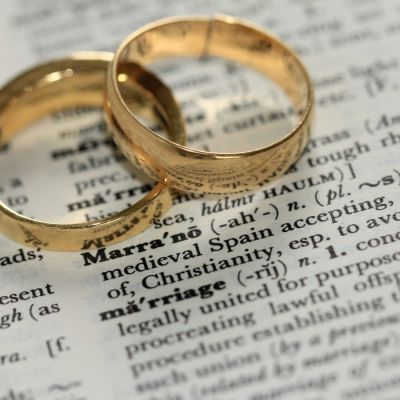 Two golden-coloured band rings atop a dictionary page zoomed in on the word ‘marriage’. Above that, the definition of the word ‘marrano’.