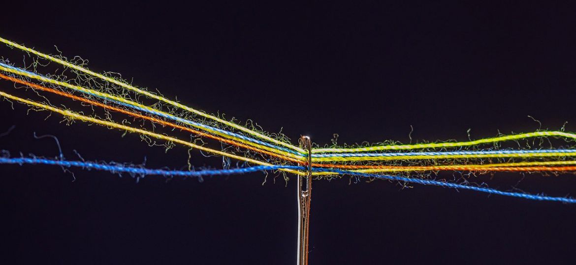 A photograph of the eye of a needle and many differently coloured threads passing through it, on a black background.
