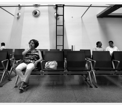 Black and white image of a woman sitting in what seems like a waiting area. Three people can be seen sitting behind her