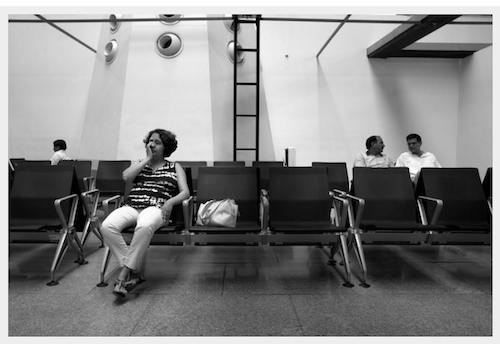 Black and white image of a woman sitting in what seems like a waiting area. Three people can be seen sitting behind her