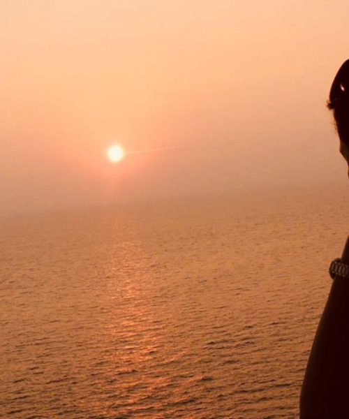 एक लड़की की तस्वीर जो समुद्र के किनारे ढलते सूरज को देख रही है
